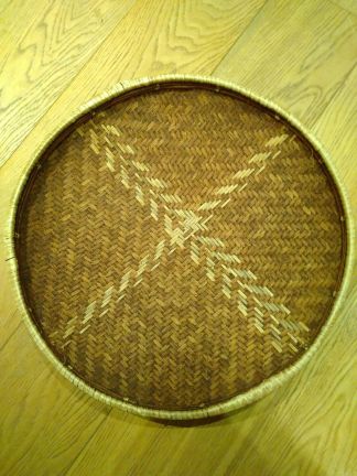 An intricately woven round, flat reed basket. The rim is finished with a lighter colour of reed. the interior is made of darker reeds, with two chevron patterns of lighter redds crossing in the centre to make a pattern like the letter X. Own photo.